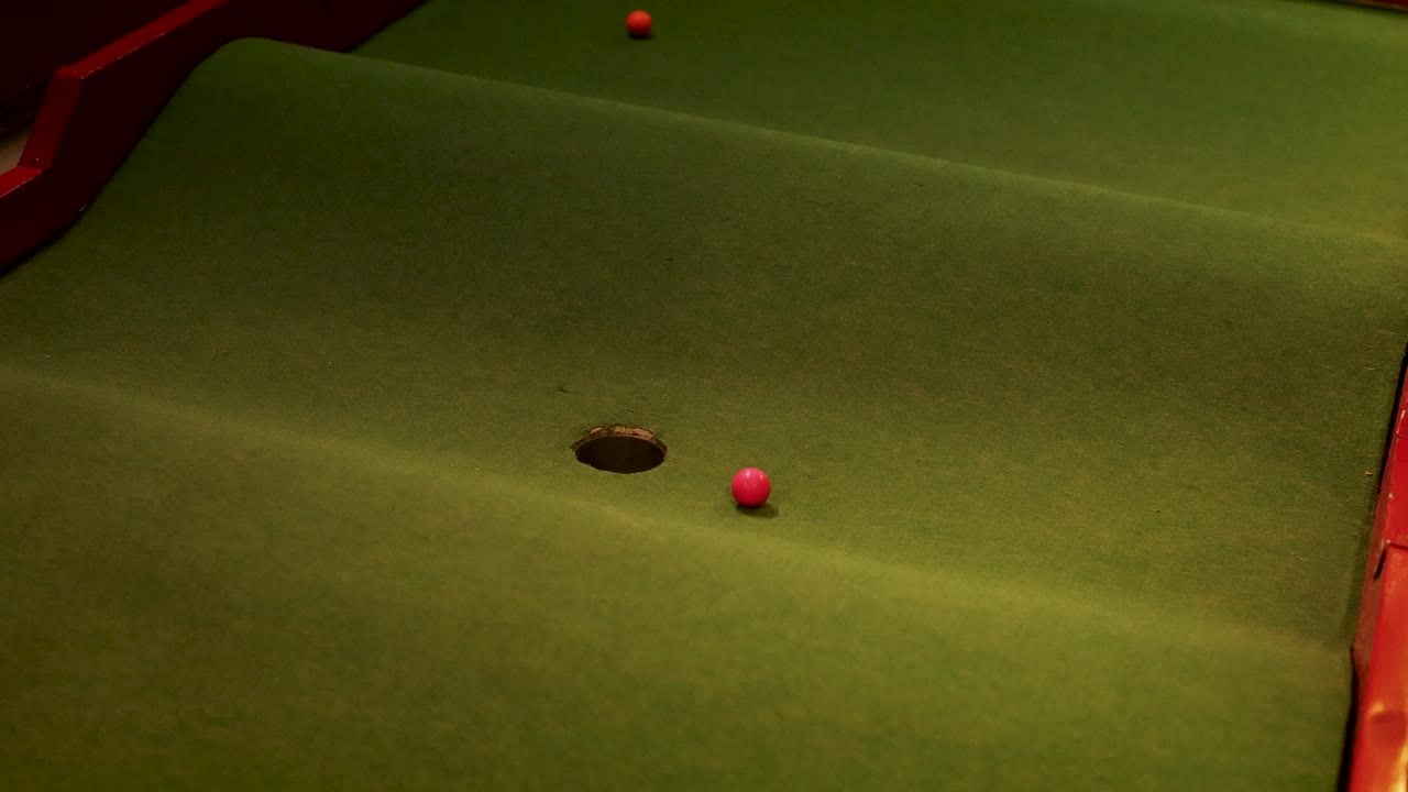 Une balle de golf roule d'avant en arrière sur un parcours miniature, finissant par tomber dans le trou sous un éclairage tamisé.