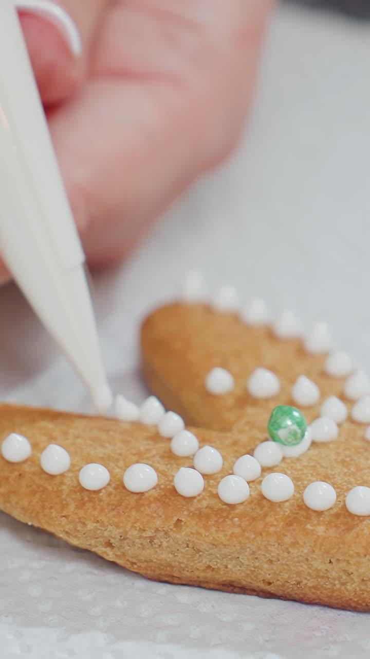 primer plano de una galleta en forma de amor decorada con puntos blancos de glaseado y una perla verde, colocada en papel de textura blanca, mano con uñas cuidadosamente cuidadas aplica cuidadosamente el glaseado con una bolsa de tubería