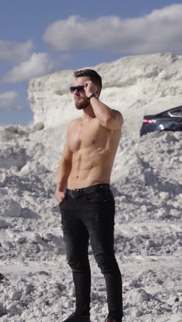 shirtless athlete on white canyon. portrait of handsome man with trained body in the mountains under the blue sky. black car on the hill at sunny day. vertical video