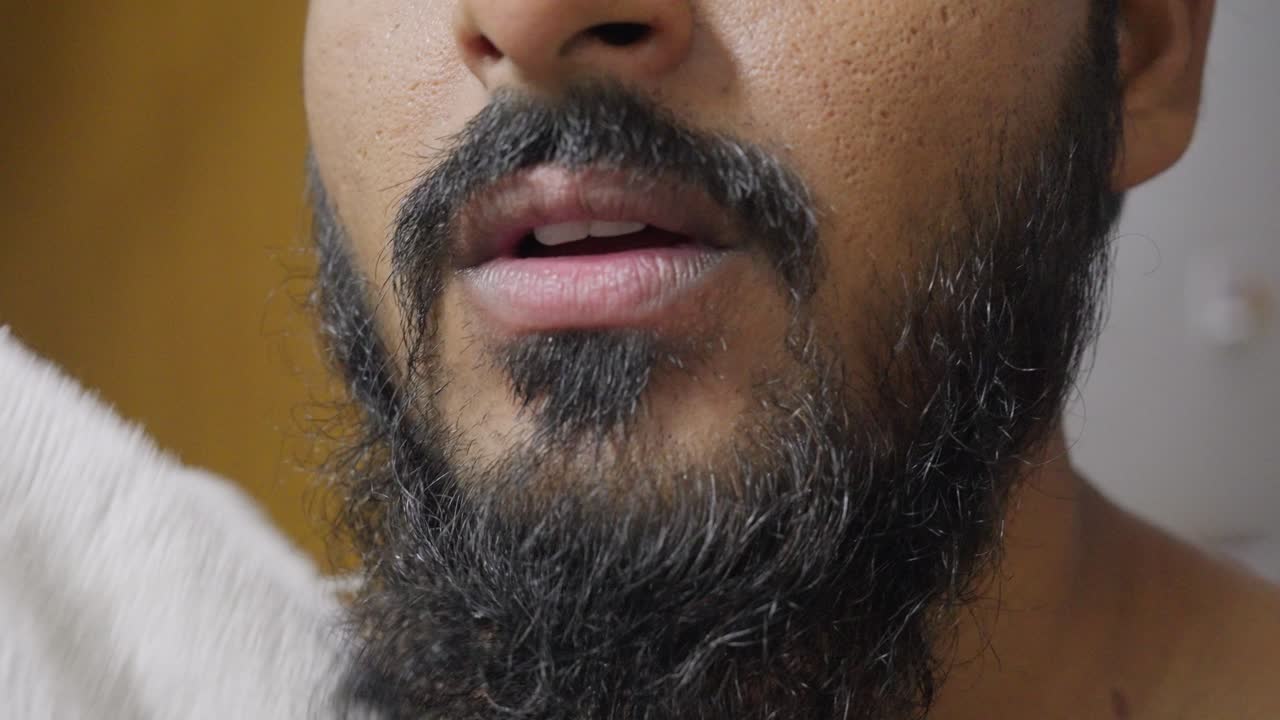 A close-up slow-motion footage of a South Asian man drying his face and beard with a white towel after brushing his teeth with blurred background