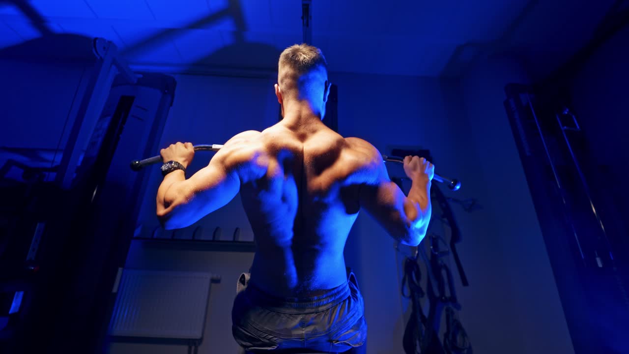 Backside view of an athlete exercising. Sportsman sitting and doing his workout on a simulator in the sports center. Blue light.