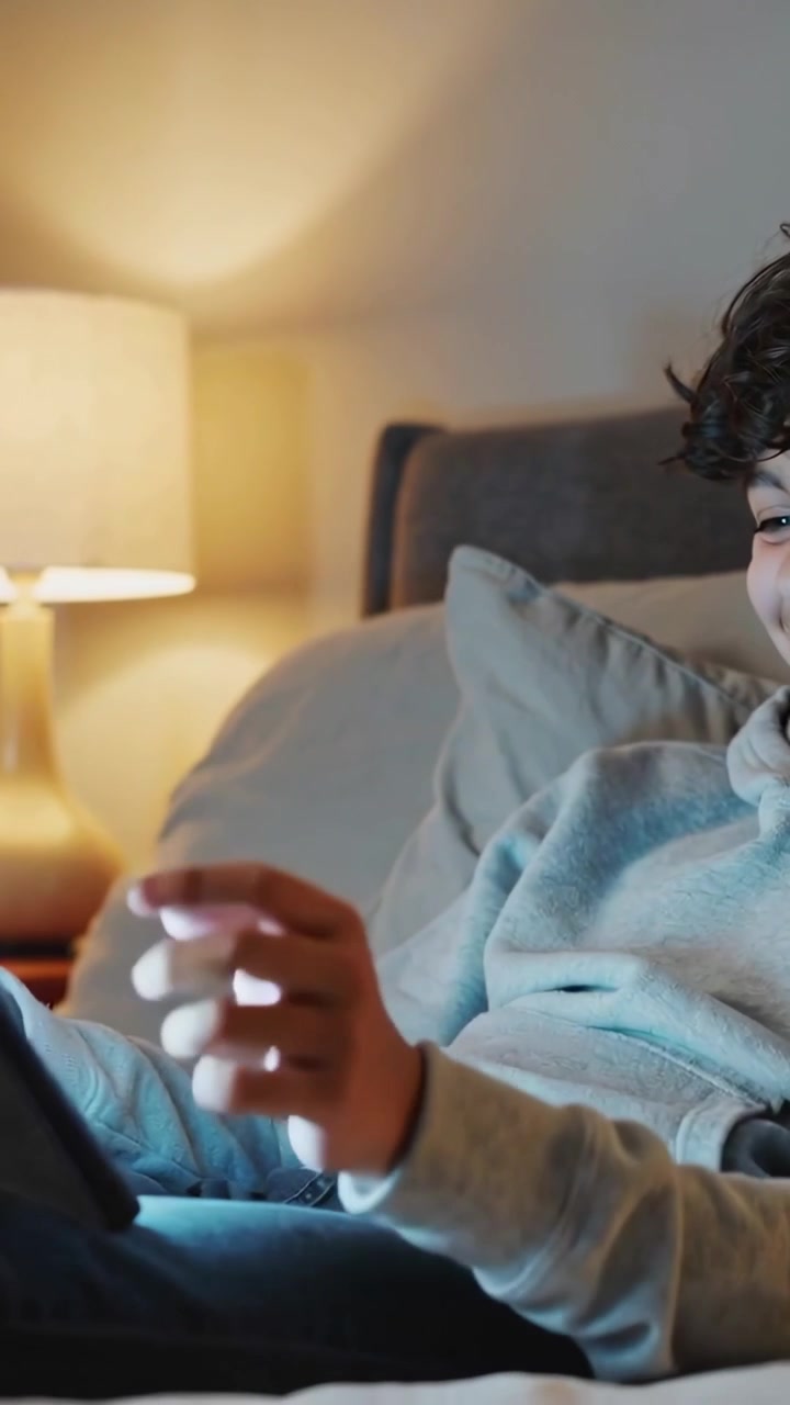 Vertical video: Looking at tablet, boy in gray hoodie tapping and swiping on bed, lamp glowing