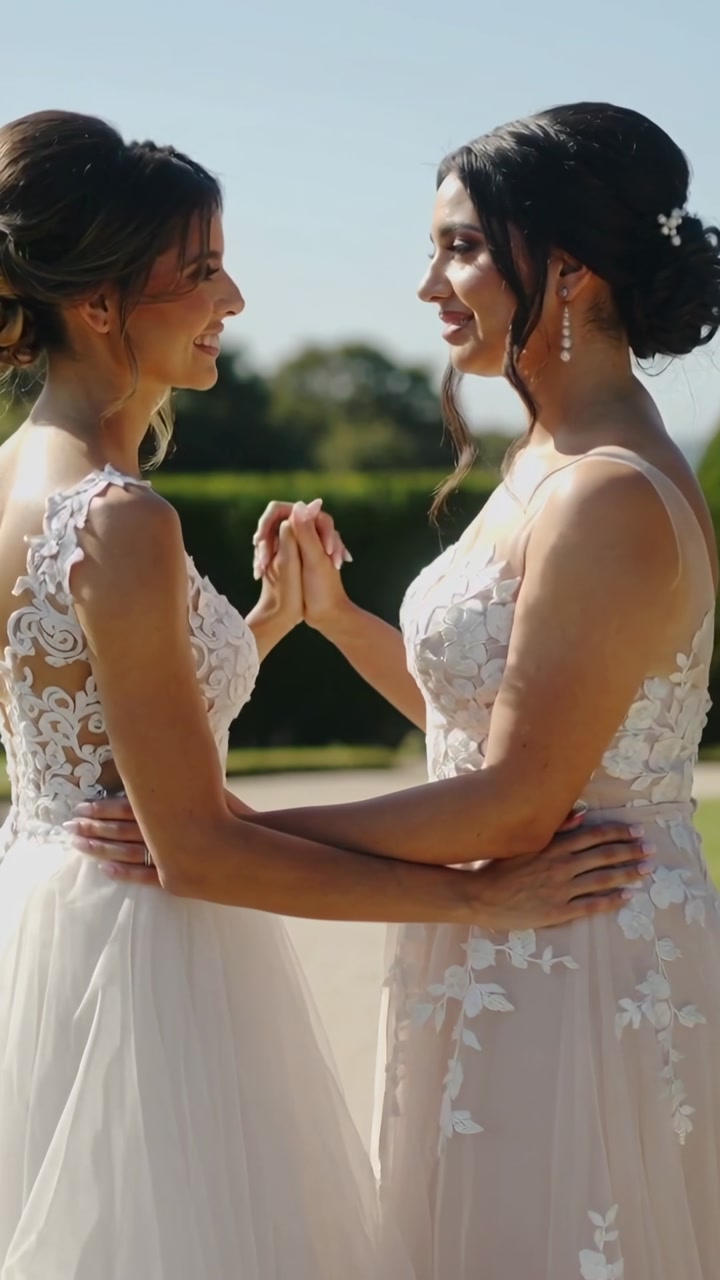 Vertical video: Holding waists, two brides exchanging gaze during first-look in garden, lace gowns