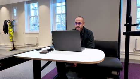 young south asian professional waving at start of webinar, sitting inside office pod, using laptop