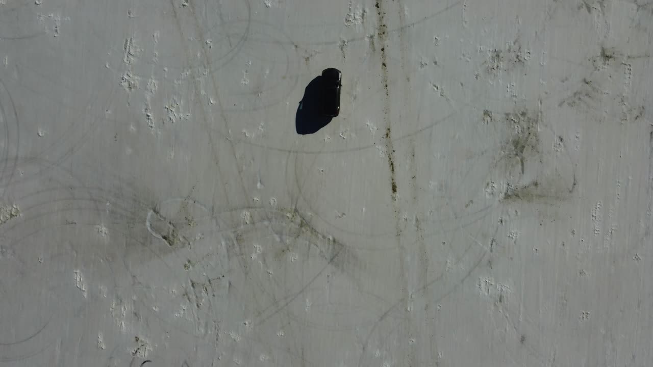 Car driving on Bonneville Salt Flats in Utah from overhead. Tire treads visible on the white lake bed speedway, shadow cast on the car providing depth. Car commercial-like 4K drone shot.