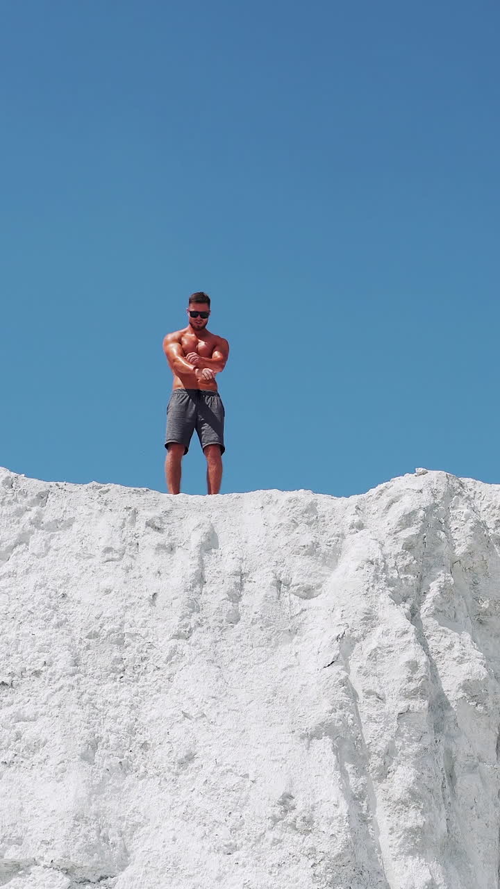 A strong man with glasses demonstrates his muscular body. Bodybuilding on the street on a white mountain against the blue sky. Vertical video