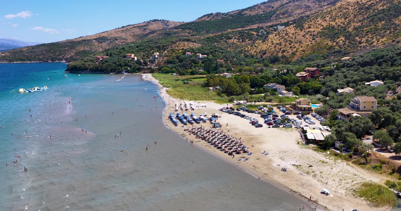 Volo di drone sopra la spiaggia di Kalamaki Apraos a Corfù, Grecia, che cattura la costa sabbiosa e poco profonda, il mare turchese e limpido e le rigogliose colline verdi