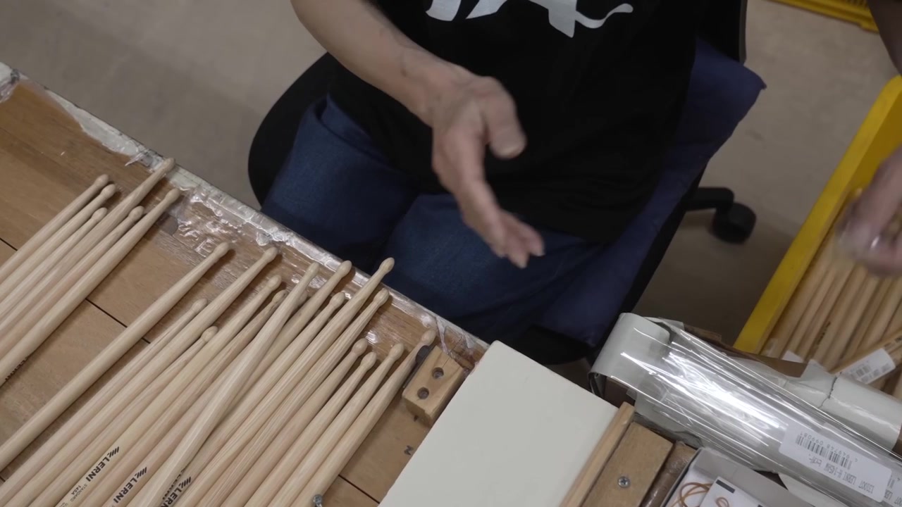 Factory worker arranging printed wooden drumsticks into a packing jig with clear plastic sleeves at a musical instrument workshop