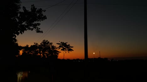 The-sun-is-setting,-casting-a-golden-glow-on-the-silhouette-of-a-tree,-while-a-gas-chimney-burns-in-the-background