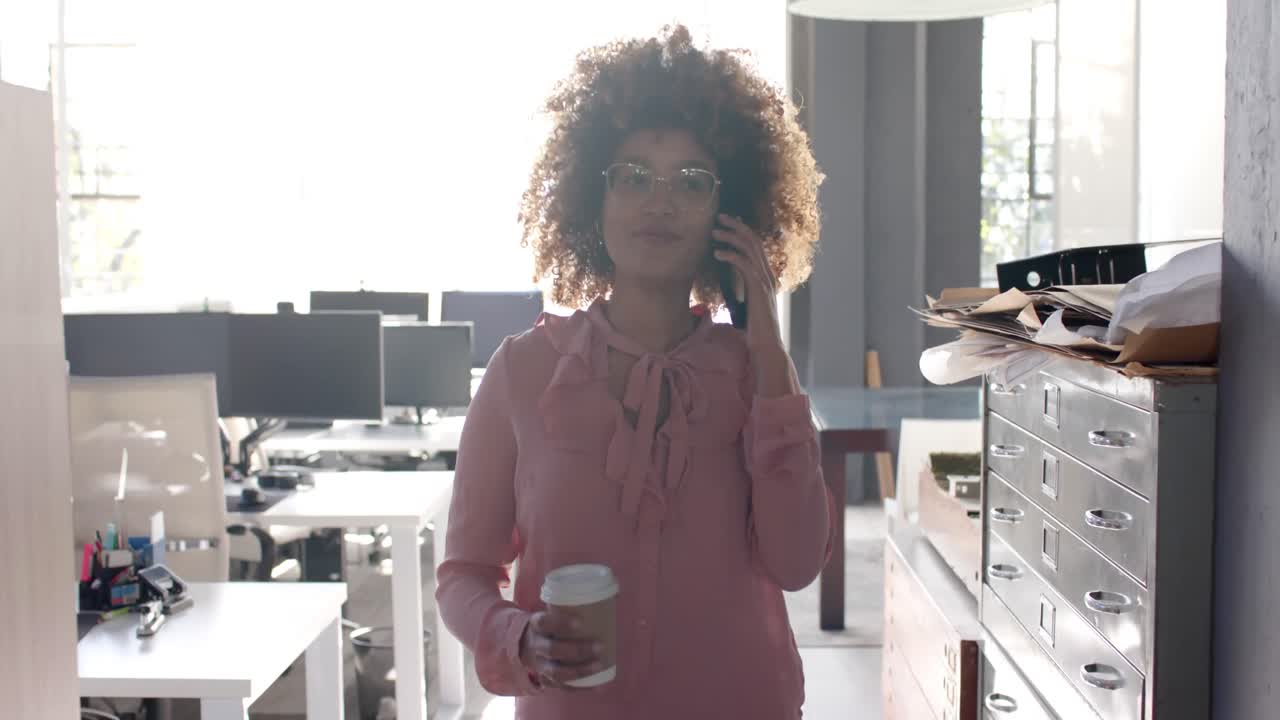 Happy biracial casual businesswoman walking through office talking on smartphone, slow motion