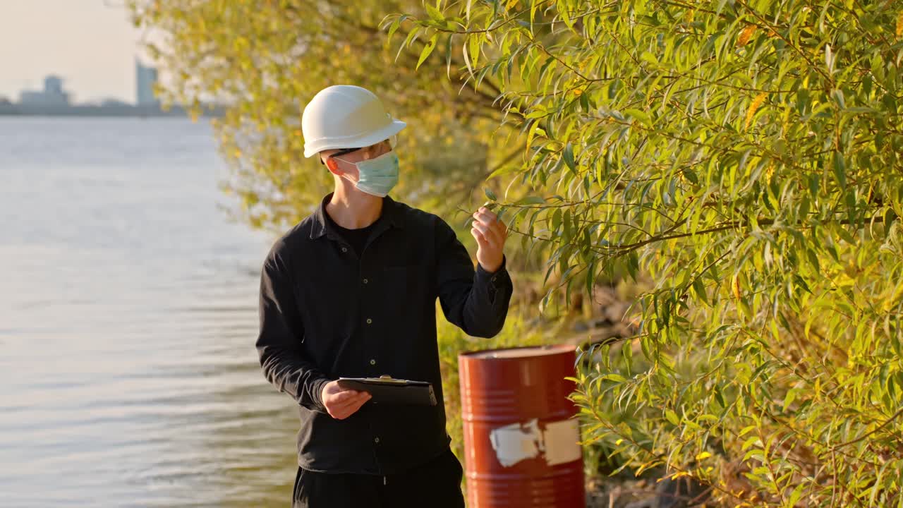 Scientist In PPE Suit, Hard Hat, And Face Mask Writing Down His Evaluation After Inspecting The Trees On The Banks Of River. - wide shot