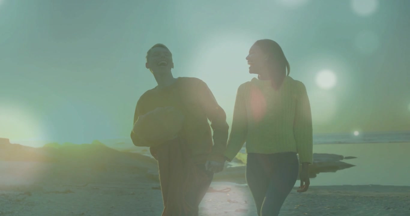 Low sun making lens flare couple holding hands walking coast male holding ball showing travel mood
