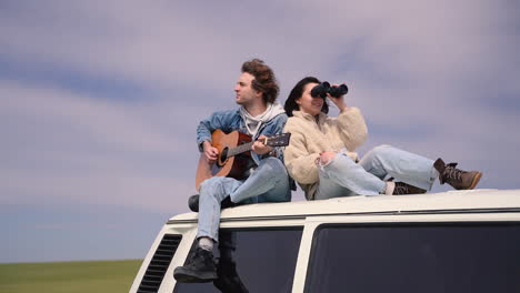 a young boy plays the guitar and a young girl looks around with a pair of binoculars on the roof of a caravan