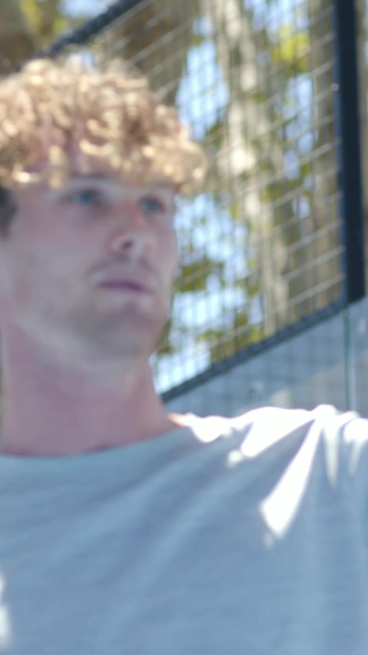 Vertical video: Reacting man in grey tee holding paddle at fence warming up, camera panning
