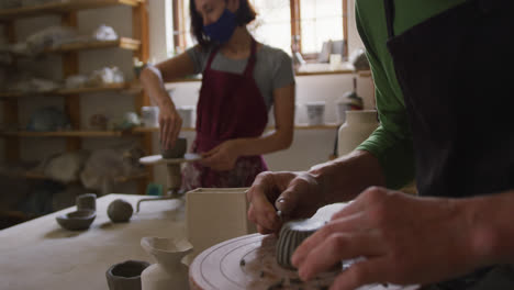 male potter wearing face mask and apron using ribbon tool to create design on pot at pottery studio