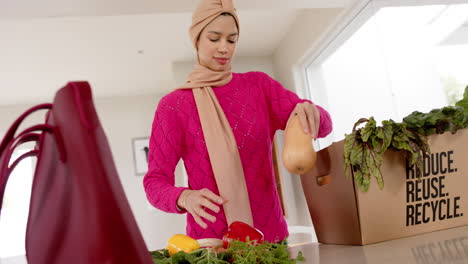 Happy-biracial-woman-in-hijab-with-grocery-shopping-in-kitchen-at-home-with-copy-space,-slow-motion