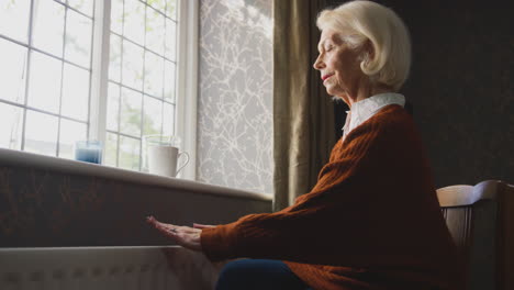 senior woman with hot drink trying to keep warm by radiator at home in cost of living energy crisis