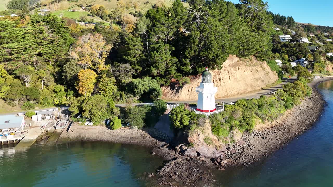 Drone footage captures Akaroa's scenic lighthouse, lush greenery, and coastal landscape in bright daylight, showcasing natural beauty and tranquility