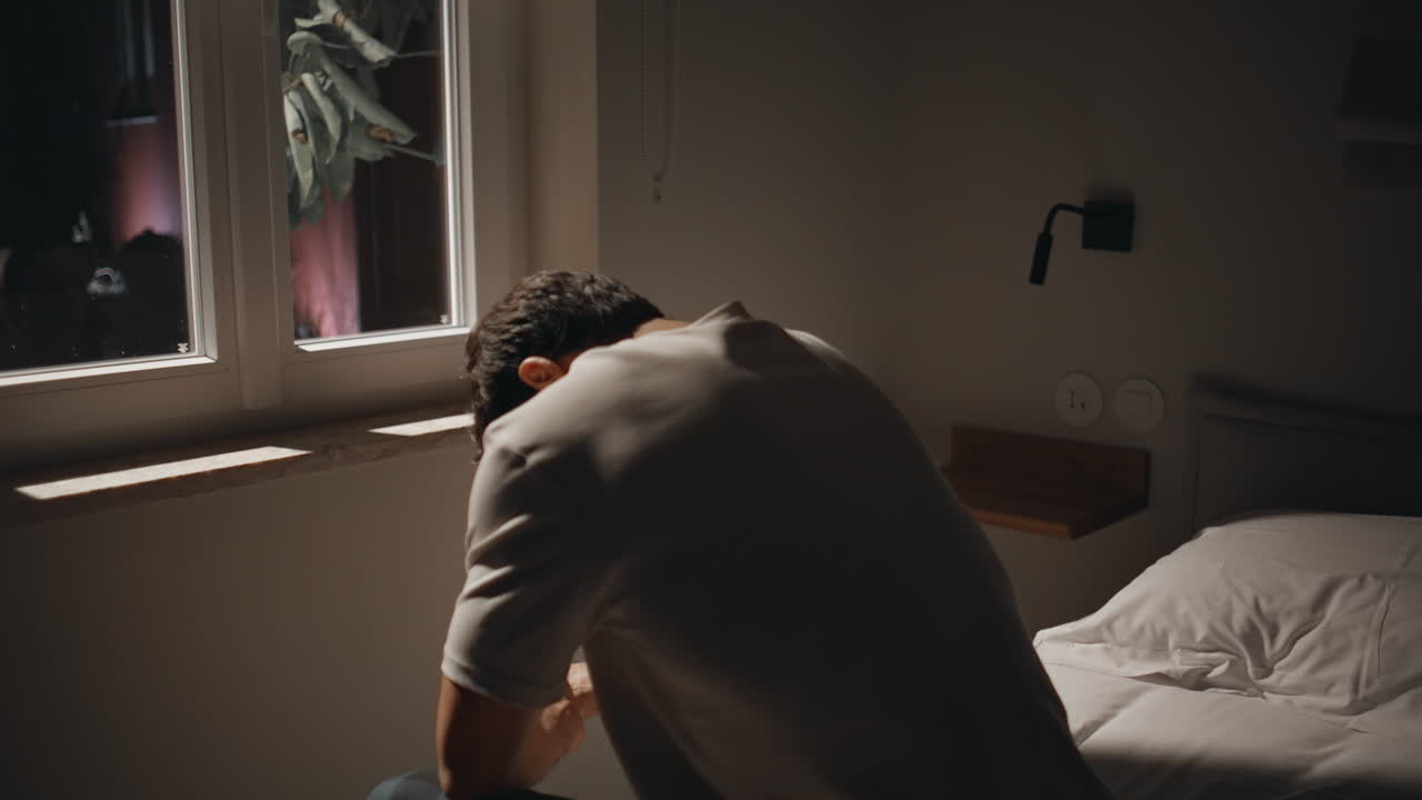 Sorrowful man sitting troubled at night bedroom reflected in dark window closeup