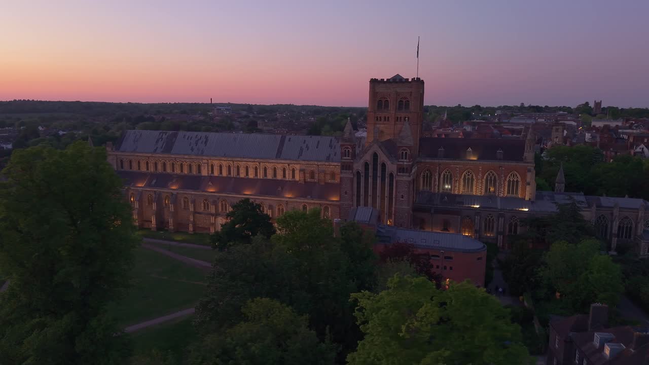 Aerial telephoto drone orbit captures the illuminated St Albans Cathedral at night, highlighting the architecture and details while keeping the city skyline in view