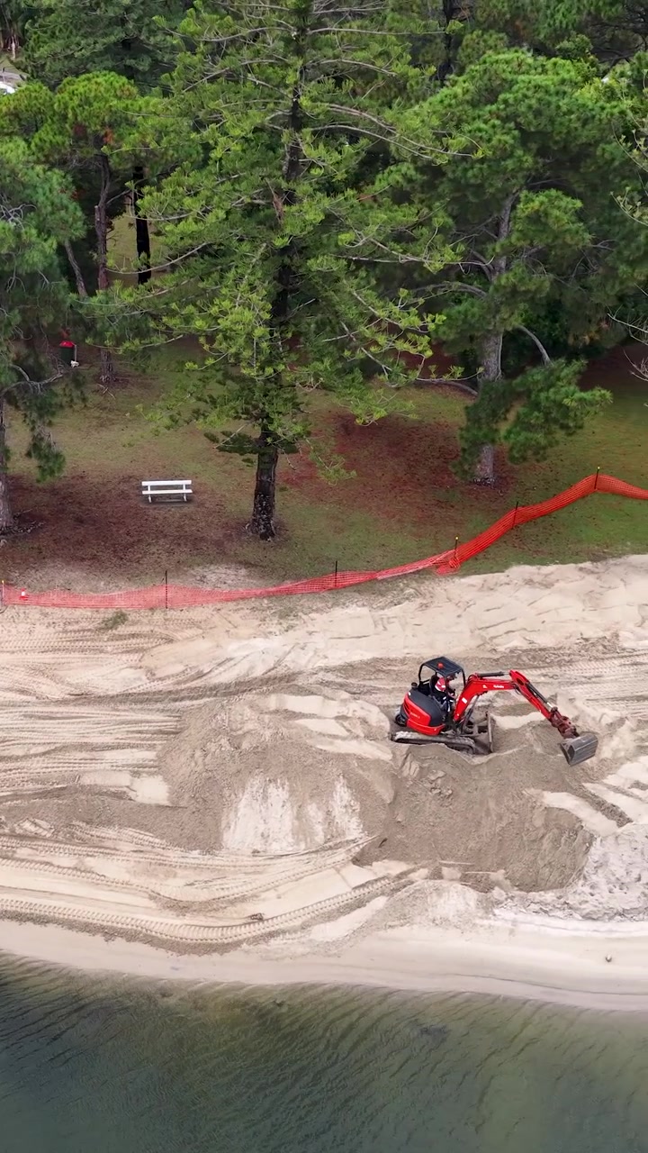 Ripresa aerea dall'alto di macchinari pesanti che spostano sabbia durante un progetto di ripristino di una spiaggia costiera.