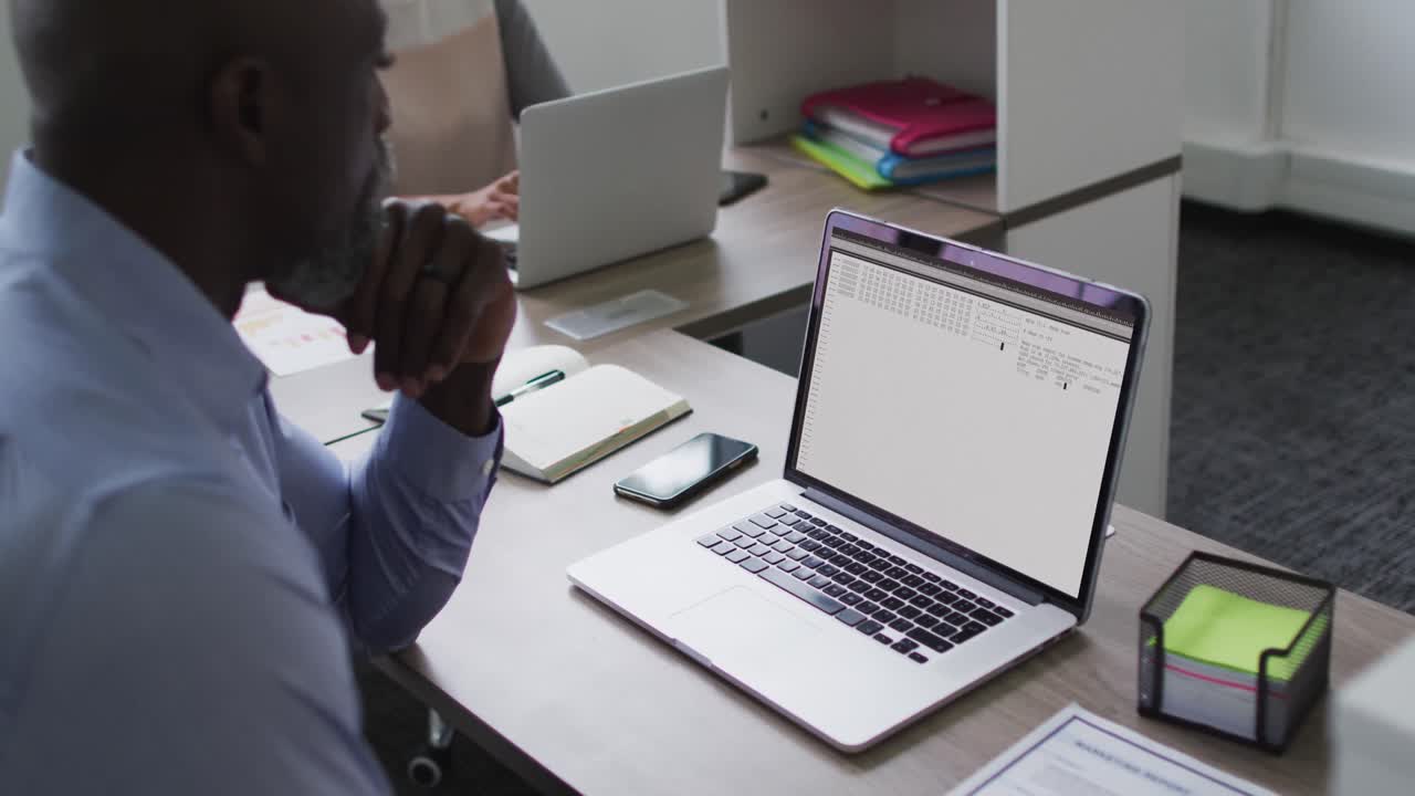 Afroamerikansk mand sidder ved skrivebord og ser på kodning og databehandling på laptopskærmen.