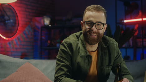 Foto-De-Retrato-Del-Joven-Feliz-Con-Gafas-Viendo-El-Canal-Deportivo-En-La-Televisión-Por-La-Noche,-Bebiendo-Cerveza-Y-Sonriendo