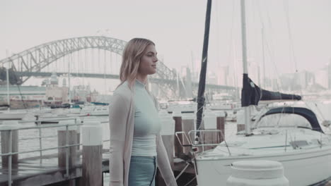 Joven-Hermosa-Mujer-Caminando-En-Un-Muelle-Con-Yates-Marina-Y-Fondo-Del-Puente-Del-Puerto-De-Sydney-En-Cámara-Lenta-Positiva-Y-Pacífica