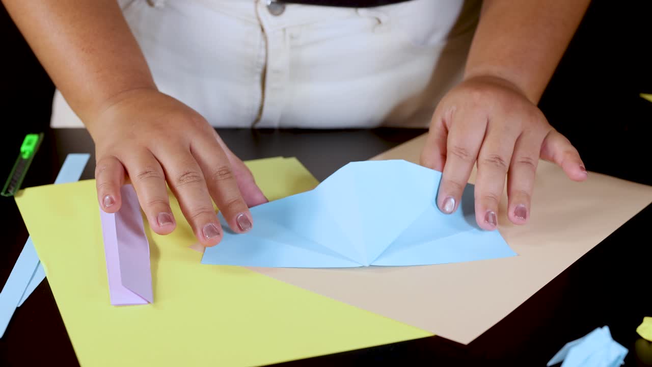 Person folds blue paper on desk under bright lighting, demonstrating step-by-step origami technique