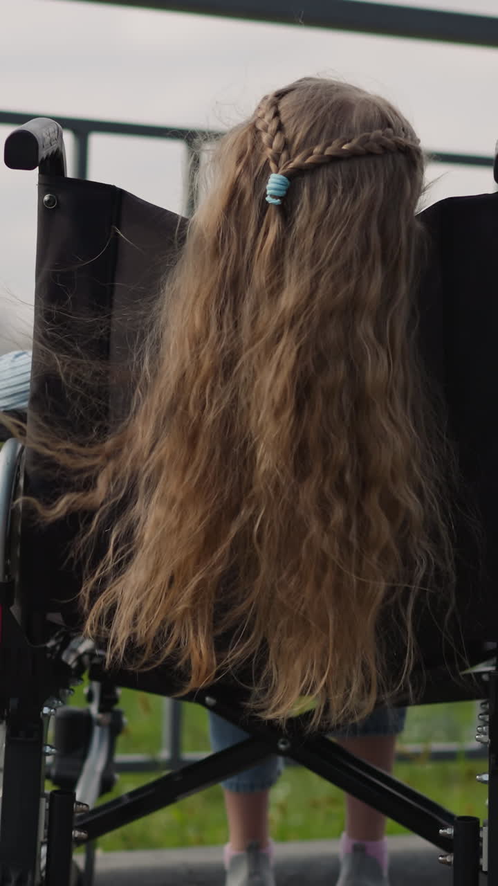 el largo cabello rojo de una niña en silla de ruedas ondas en el viento. niña de escuela junior pelirroja con discapacidad disfruta del panorama de la ciudad en el punto de vista de la parte trasera