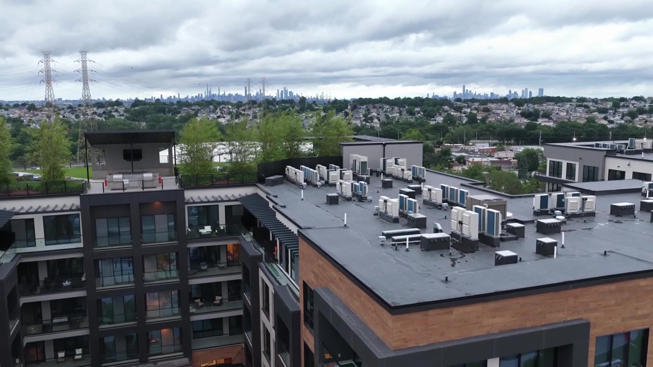 HVAC-enheder på taget pryder et moderne lejlighedskompleks i Belleville, New Jersey, med glasbalkoner og murstensfacade, der indrammer en central gårdhave, mens Manhattans skyline toner frem i det fjerne. Stigende vidvinkeloptagelse med drone.