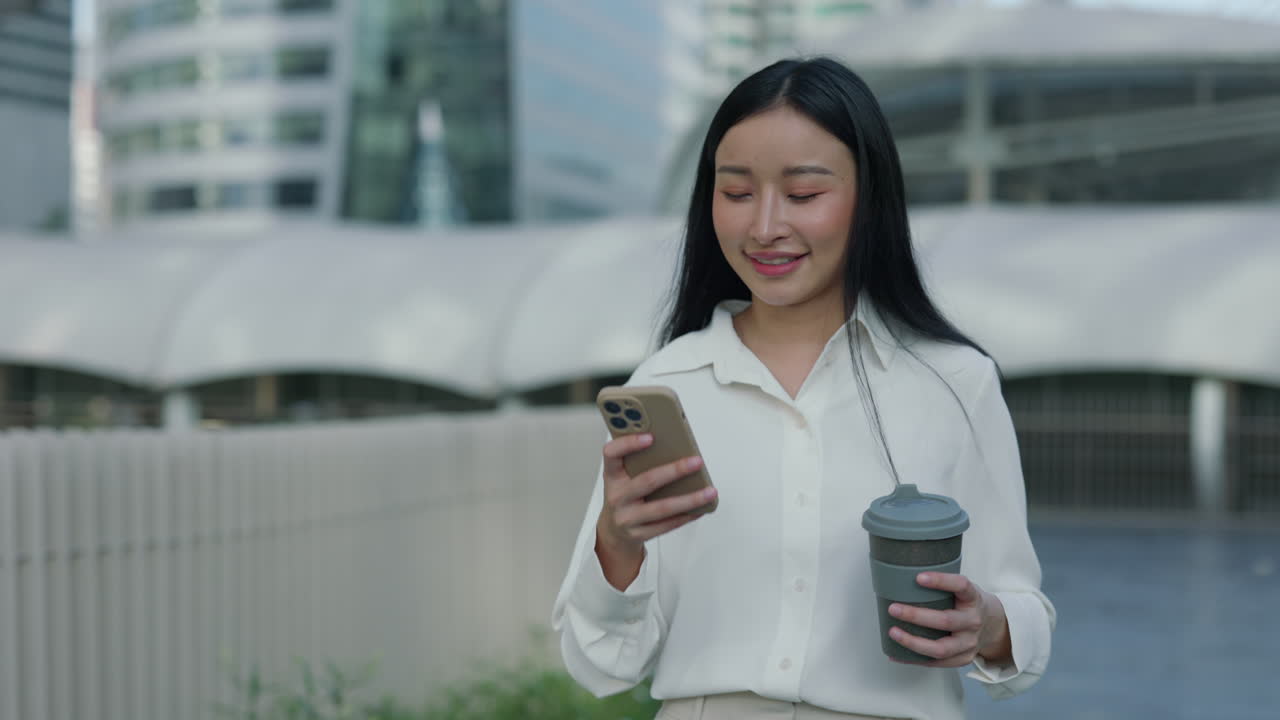 Smiling Asian woman holding coffee cup and looking at smartphone in urban setting