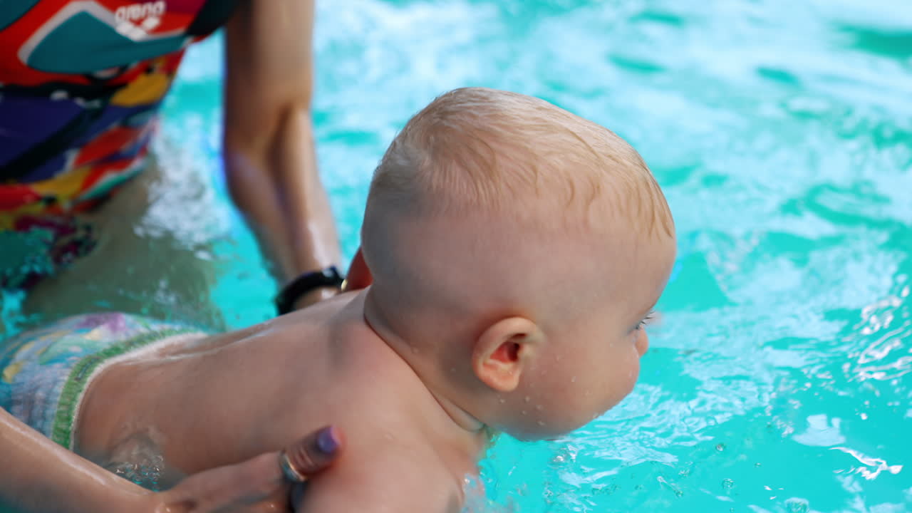 Cute Caucasian baby boy moving in the pool. Female hands hold the child splashing water in the swimming pool.