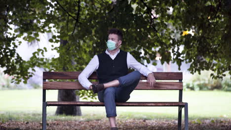 An-elegant-young-man-in-a-park-wearing-a-black-waistcoat-and-white-shirt-and-a-green-protective-Covid-19-mask,-sitting-on-a-park-bench,-browsing-his-phone-then-putting-it-into-his-pocket,-static-4k