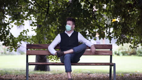 Un-Joven-Elegante-En-Un-Parque-Con-Un-Chaleco-Negro-Y-Una-Camisa-Blanca-Y-Una-Máscara-Protectora-Verde-Covid-19,-Sentado-En-Un-Banco-Del-Parque,-Mirando-Su-Teléfono-Y-Luego-Metiéndolo-En-El-Bolsillo,-Estático-4k