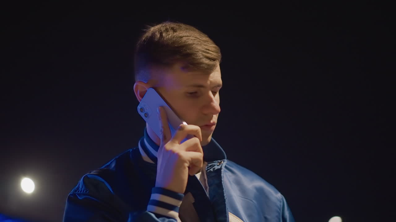 밤 고속도로에서 휴대폰을 사용하는 젊은 남성, 헤드라이트 아래 파란색 재킷, 내비게이션을 확인하고 발송 담당자와 픽업을 조율하며, 네온 불빛이 반사되는 가운데 도로 옆에서 꾸준한 속도로 걷는 모습