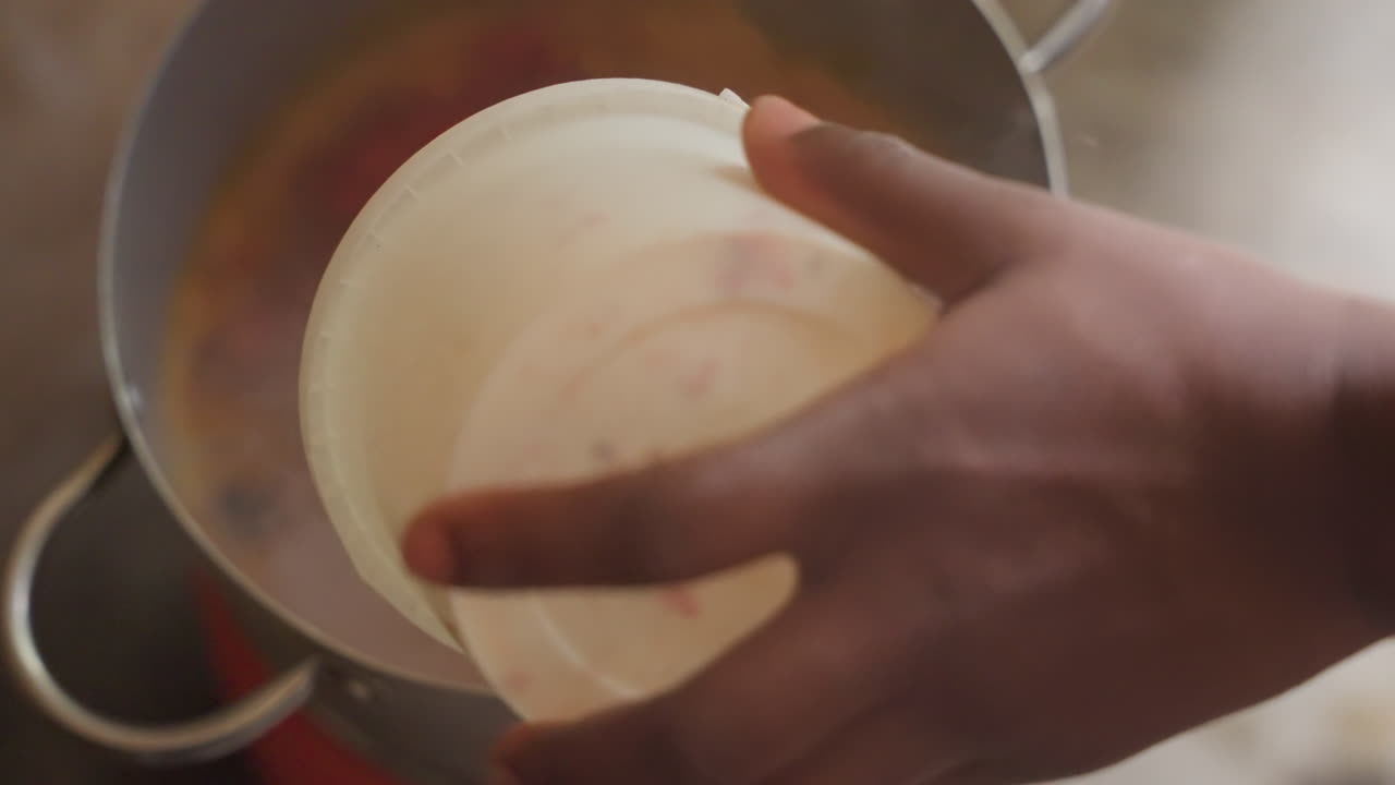 Hand pouring spice into simmering pot, colorful oil ring and rising steam, quick cloud of vapor envelops pan, rustic bowl and utensil visible, intimate streetkitchen seasoning scene