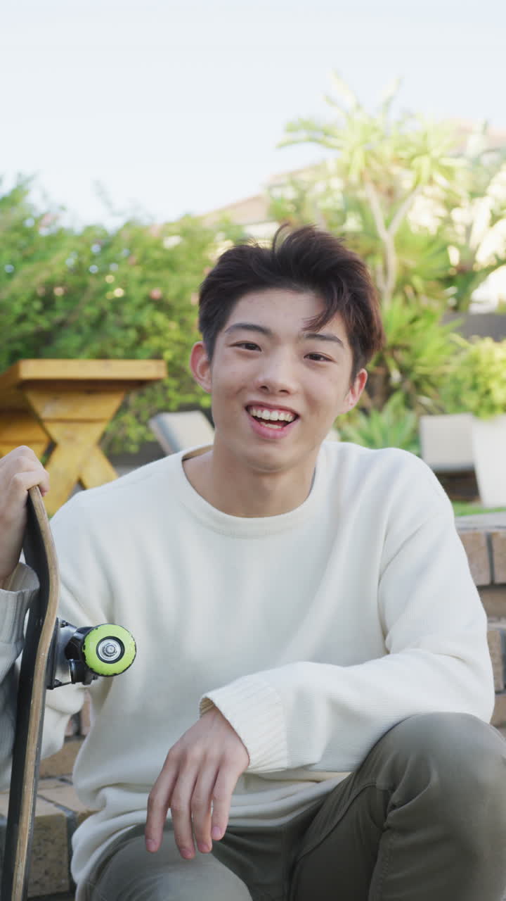 정원에서 스케이트보드와 함께 미소 짓는 아시아 남성 십대 초상화의 수직 비디오