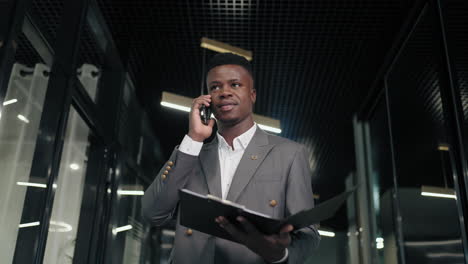 successful black business man talks to smartphone in office interior. african american businessman walks down corridor and calls on her mobile phone. portrait of friendly man walking down corridor .