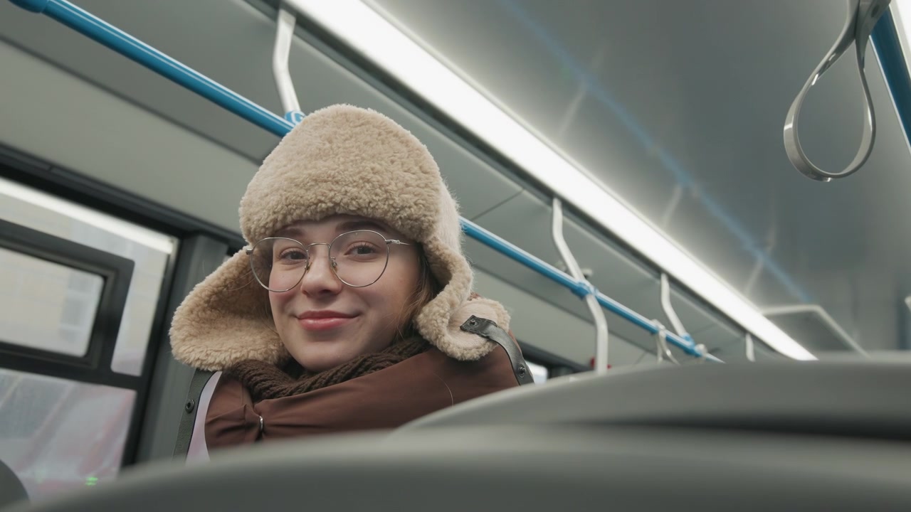 Pendolare seduto sui mezzi pubblici si china leggermente in avanti, afferrando il maniglione superiore con una mano, indossando un cappello di maglia beige con paraorecchie, occhiali e un piumino marrone, mostrando un'espressione calma e rilassata.