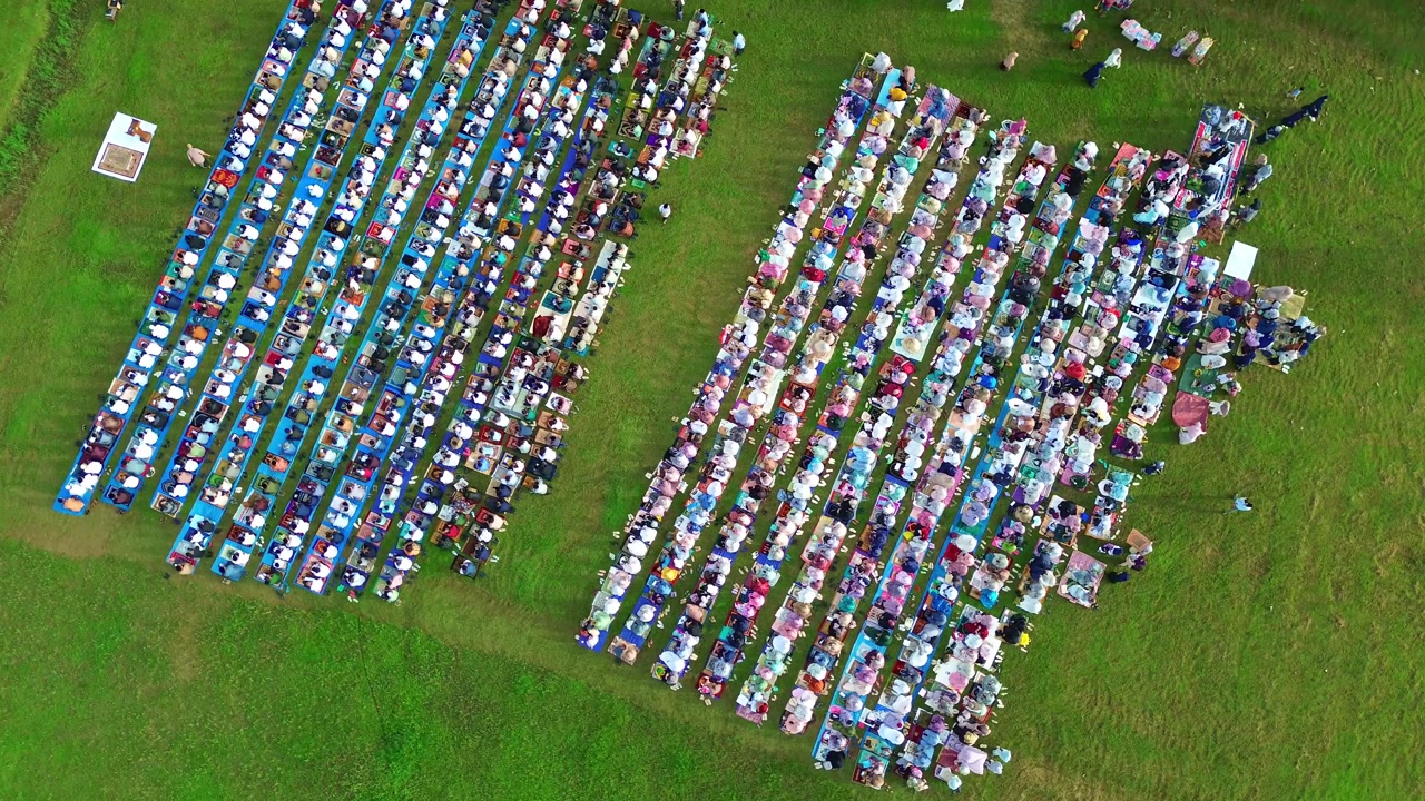 Aerial view of Eid al-Fitr prayer with Muslims gathered in neat rows on a wide green field under daylight.