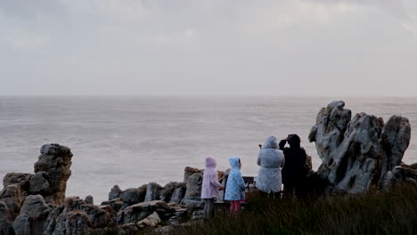 Zeitlupenaufnahme-Einer-Familie-Von-Hinten-Auf-Einer-Klippe-An-Der-Küste,-Die-Zusieht,-Wie-Eine-Welle-Gegen-Die-Felsen-Schlägt