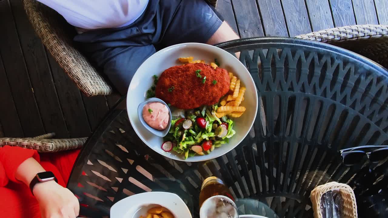 Top view of a person dining with a plate of breaded meat, fries, and a fresh salad on a glass table. Ideal for themes related to food, casual dining, and restaurant meals.