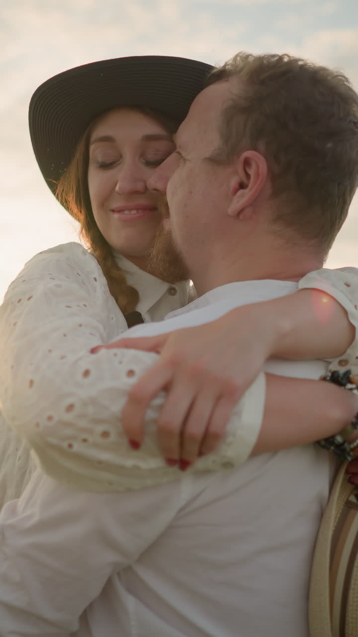 Two lovers embrace tenderly and smile in a grassy field at sunset, both dressed in white outfits. The woman wears a hat and holds another in her hand, enjoying the warmth of the golden hour