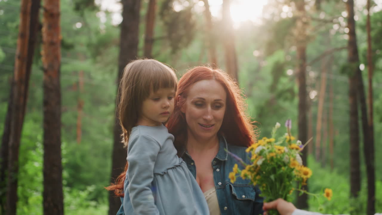 Mother with red hair carrying little girl walks through forest toward older daughter offering colorful bouquet, warm sunlight shining through trees, symbolizing family love, and nature connection