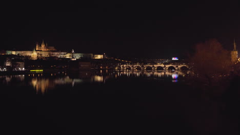 El-Castillo-De-Praga-Y-El-Puente-Charles-Sobre-El-Río-Vltava-En-El-Centro-Histórico-De-Praga,-Chequia,-Iluminado-Por-Luces-En-La-Noche,-Disparado-Desde-El-Otro-Lado-Del-Río,-Un-Muelle-Con-Farolas-Y-Rieles-De-Tranvía