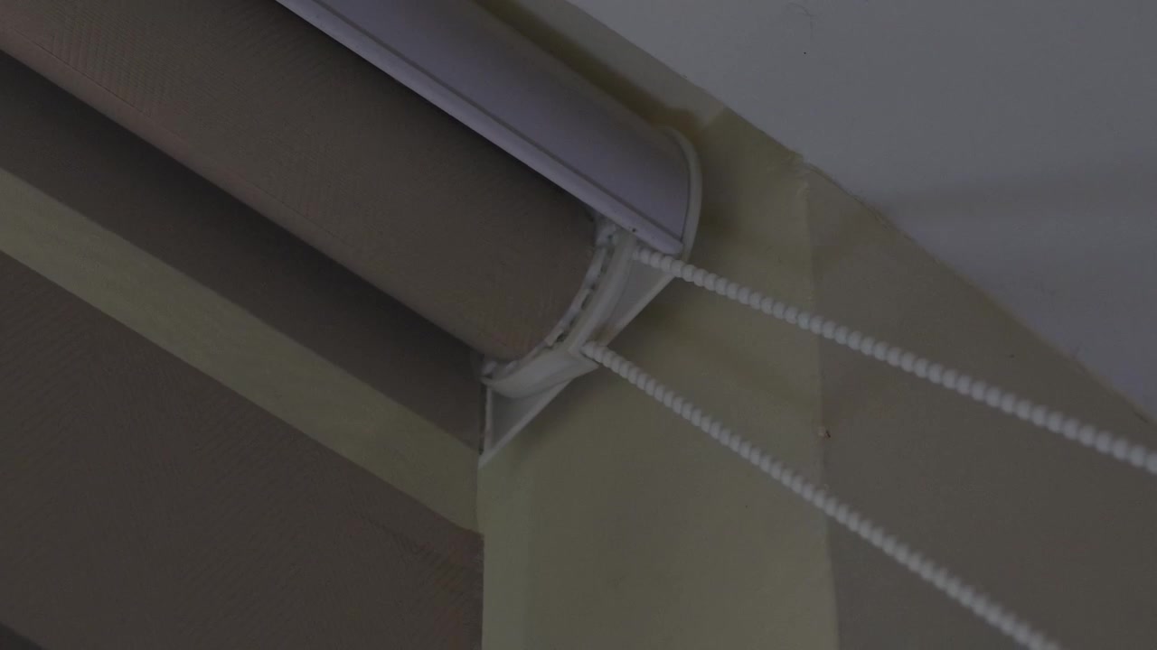 Close up shot of the top right corner of a roller blind fixture, featuring a white plastic bracket, brown rolled fabric, and a beaded white chain