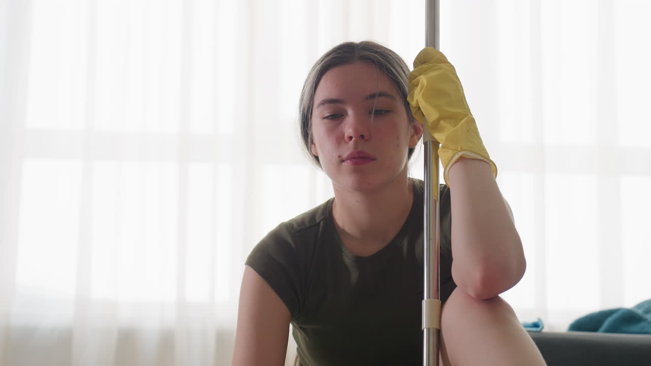 Thoughtful woman seated wearing yellow gloves resting head against mop stick adjusting herself while taking deep breath in bright living room interior with sunlight through curtain and grey sofa
