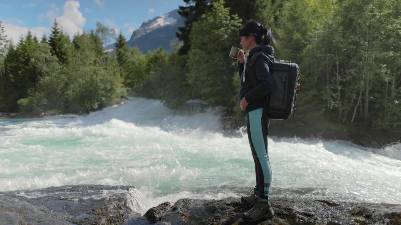 viajera con una mochila, bebiendo agua en la naturaleza en el bosque cerca de un río de montaña.