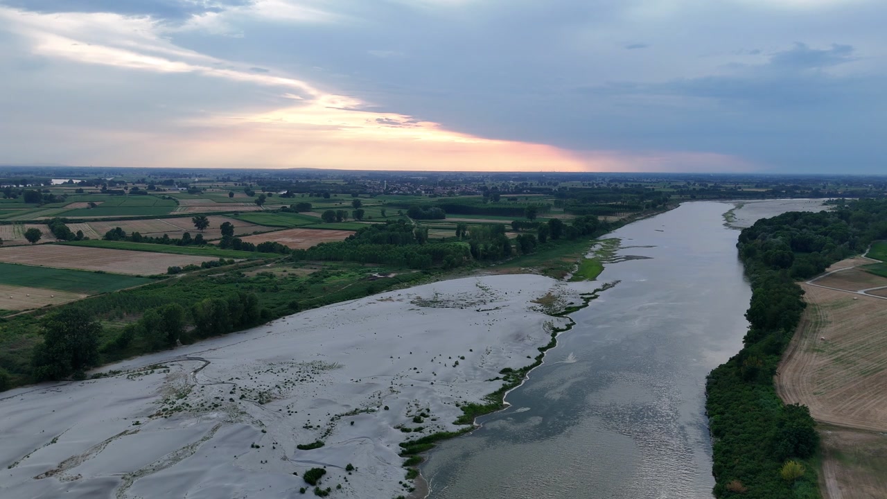 Il fiume Po mostra estesi sedimenti esposti e livelli d'acqua ridotti accanto a una pianura alluvionale coltivata vicino a Cremona, Lombardia, con sponde boscose e campi agricoli, statico.